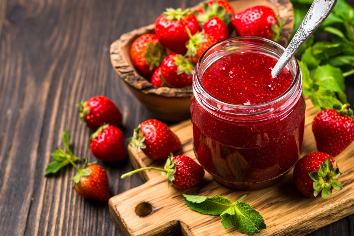 Strawberry jam in glass jar with spoon inside on wooden pad around many strawberries