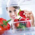 Young woman smiling and taking a fresh strawberry from refrigerator.
