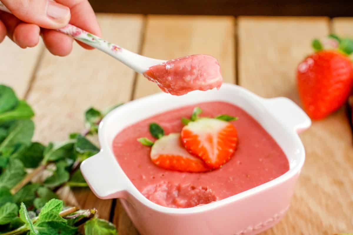 Hand holding a spoon over bowl full of strawberry pudding on a wooden table.