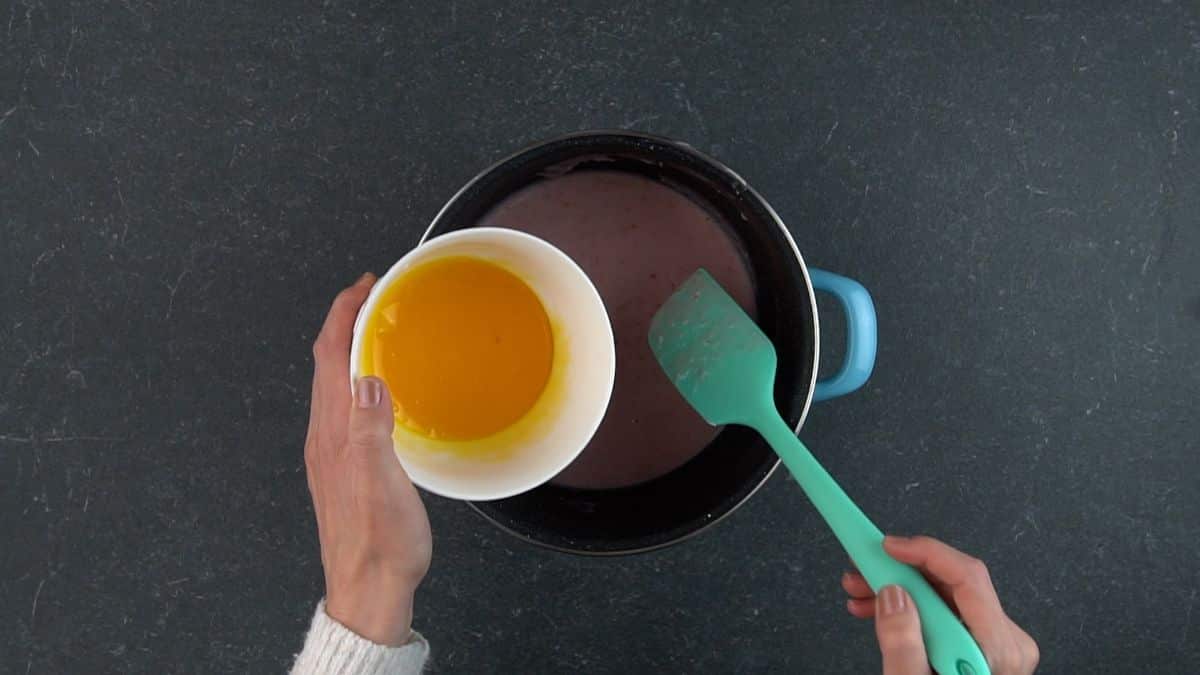 Hands stirring mix in a pot with blue spatula and holding a bowl of yolks.