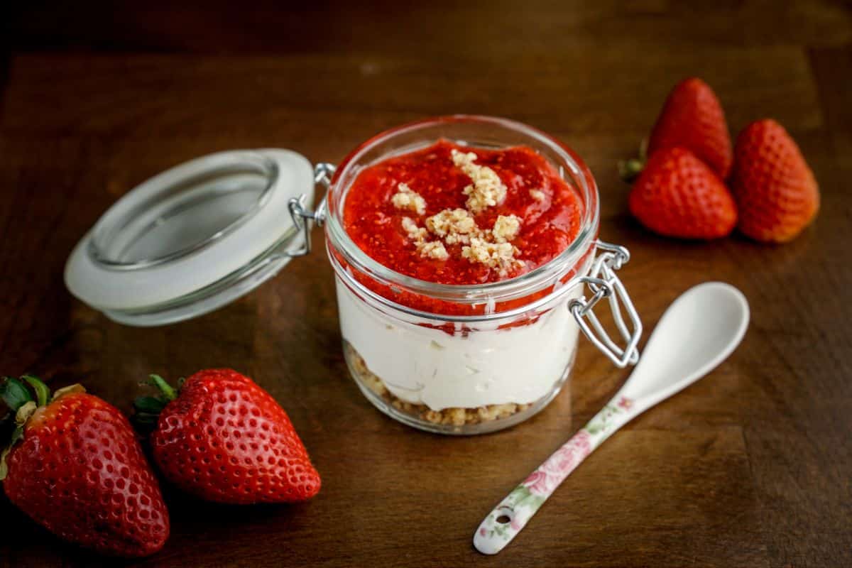 A No Bake Keto Strawberry Cheesecake Jar on a brown table next to a plastic spoon and ripe strawberries.