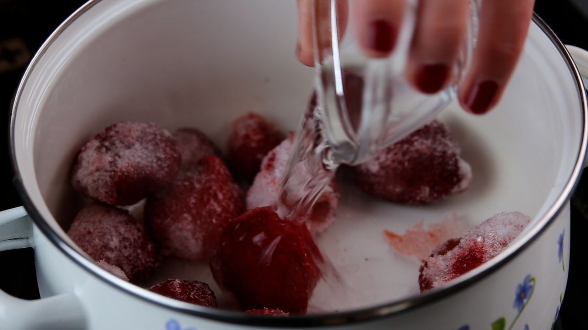 A hand pouring water in a pan with frozen strawberries.