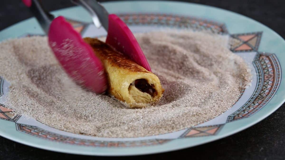 Kitchen tongs rolling a rolled piece of bread with nutella in a sugar mixutre on a plate.