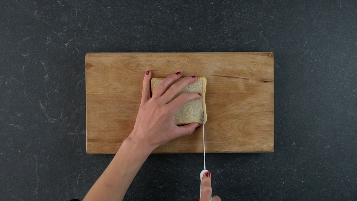 Hands cutting a crust of a slice of bread with a knife on a wooden cutting board.