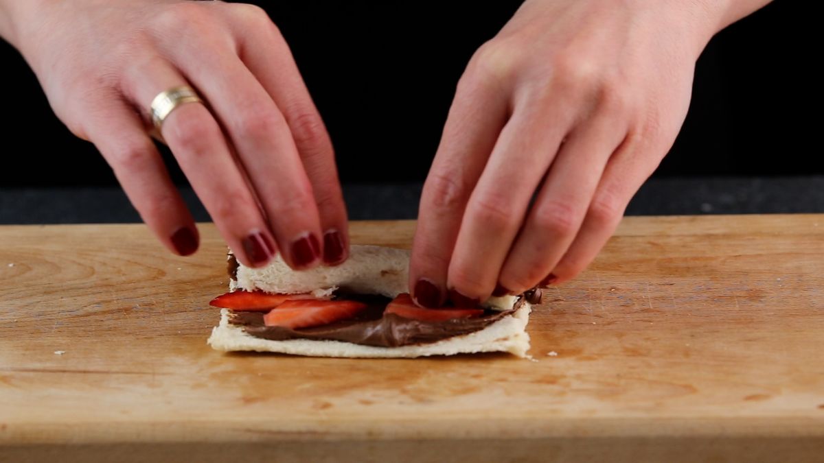 Hands adding sliced strawberries on a rolled piece of bread with nutella on a wooden cutting board.