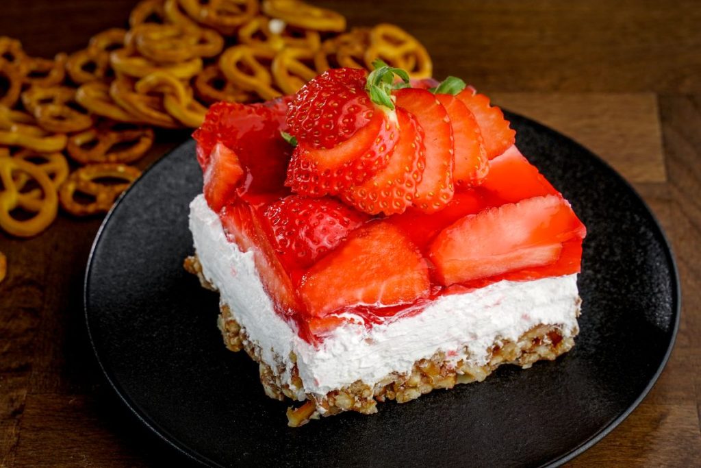 Piece of strawberry pretzel salad on a blac ktray with pretzels in the background.