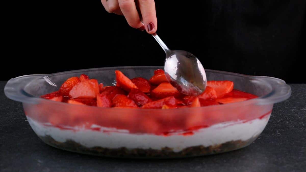 Hand with spoon spreading cutted strawberries over dish in a container.