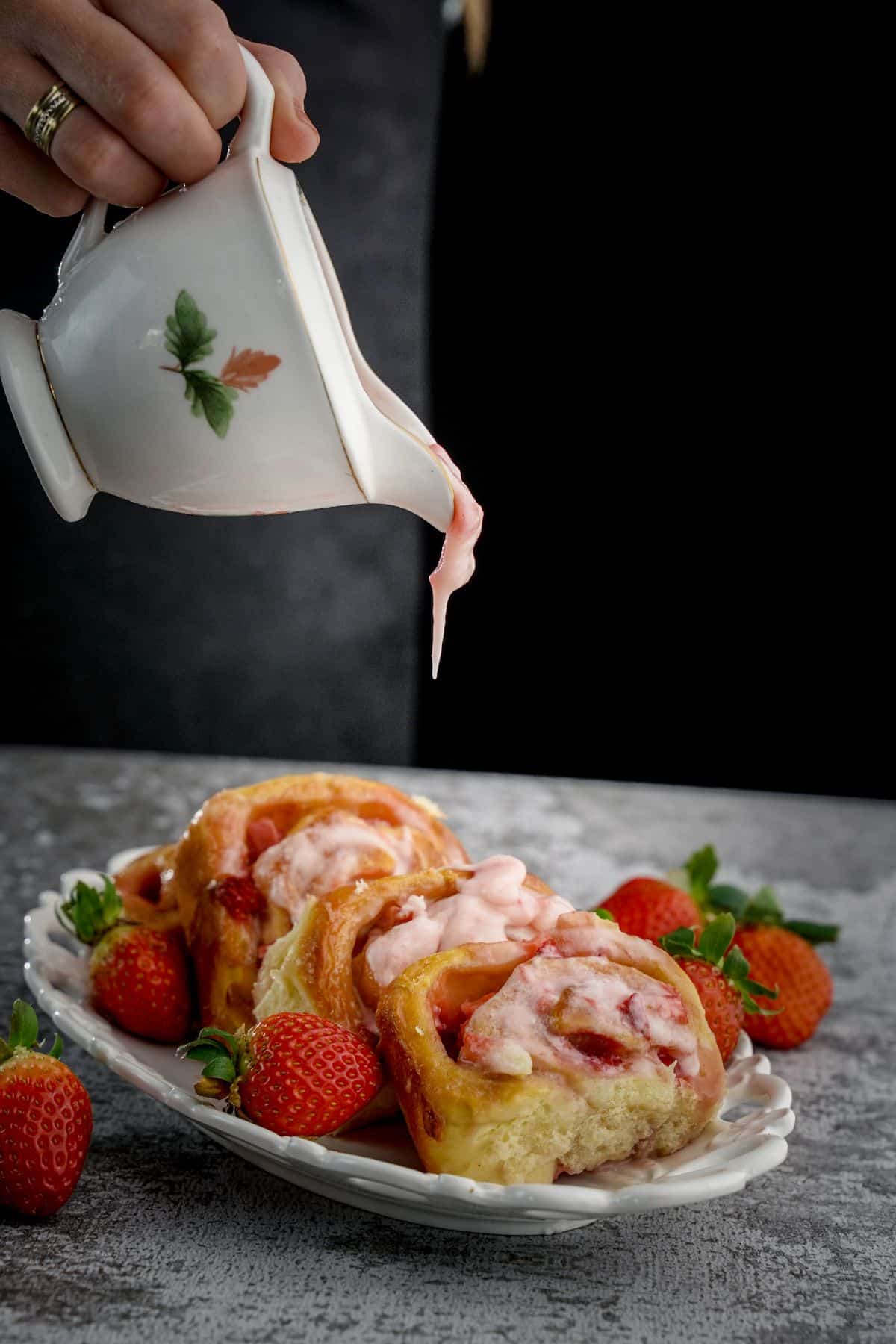 A hand pouring a strawberry frosting on Strawberries and Cream Sweet Rolls on a white tray.