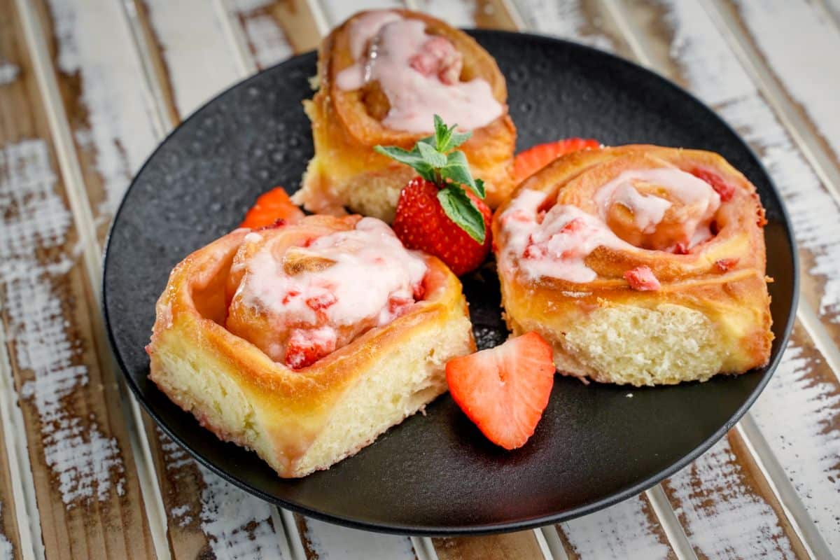 Three Strawberries and Cream Sweet Rolls on a black plate with ripe strawberries on a wooden table.