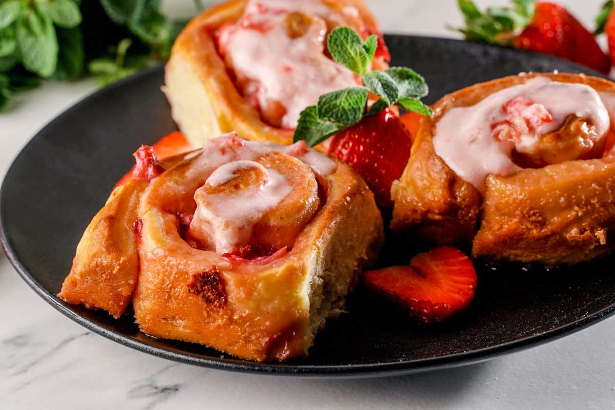 A close-up of Strawberries and Cream Sweet Rolls on a black plate with ripe strawberries.