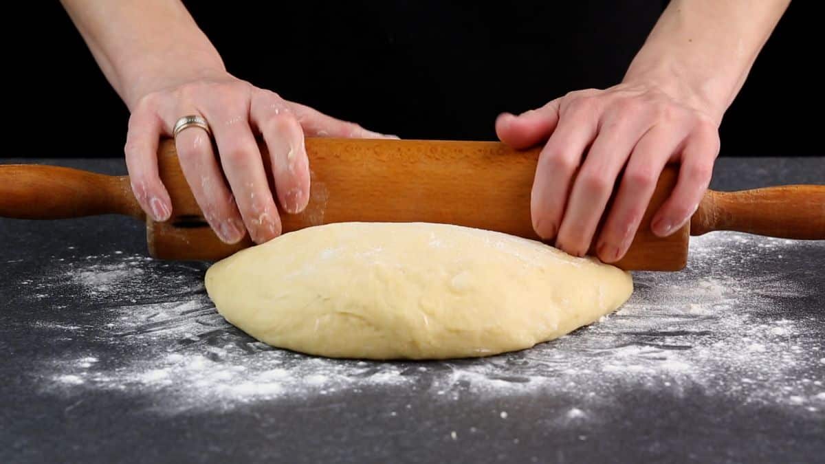 Hands rolling a dough on a floured table with a rolling pin.
