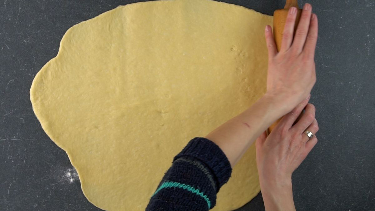 Hands rolling a dough on a floured table with a rolling pin.