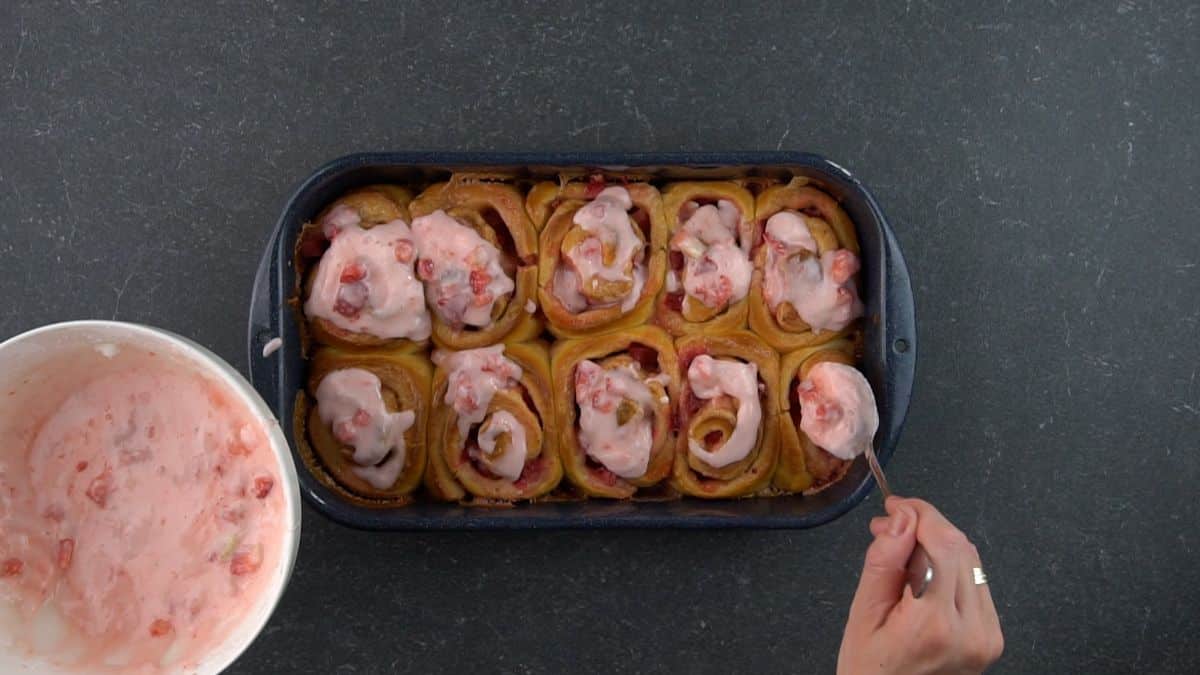A hand with a spoon drizzling a strawberry frosting over rolls in a pan.