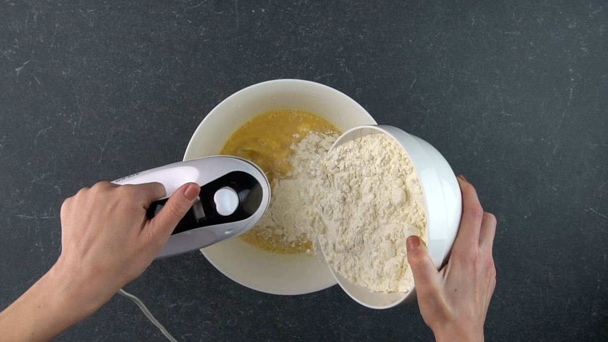 Hands puring a flour in a big bowl and mixxing with an electric mixxer.