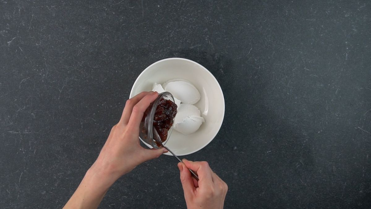 Hands scooping a strawberry jam with a scoop in a white bowl.