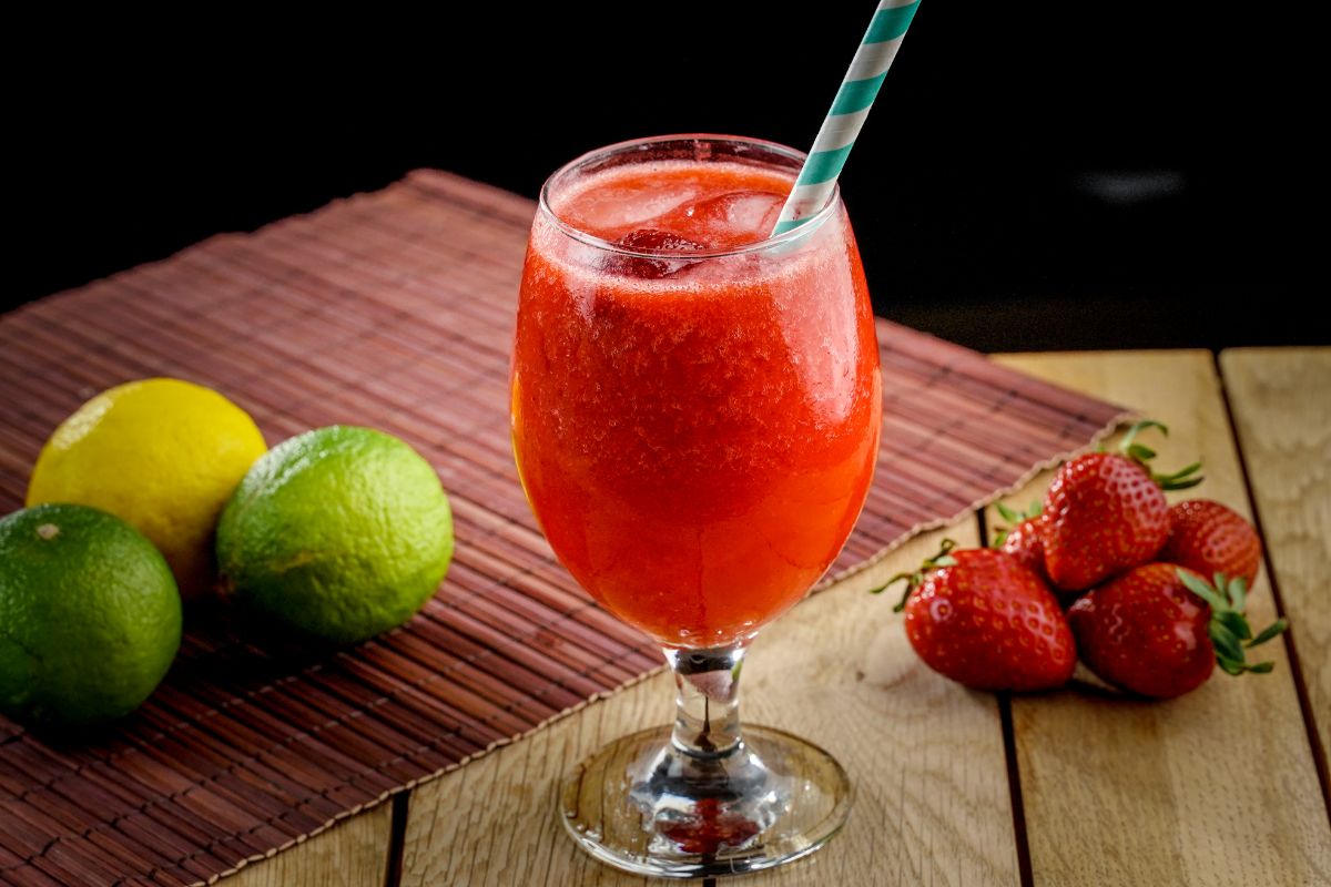Strawberry daiquiri in a glass cup with straw on the table with limes and ripe strawberries.