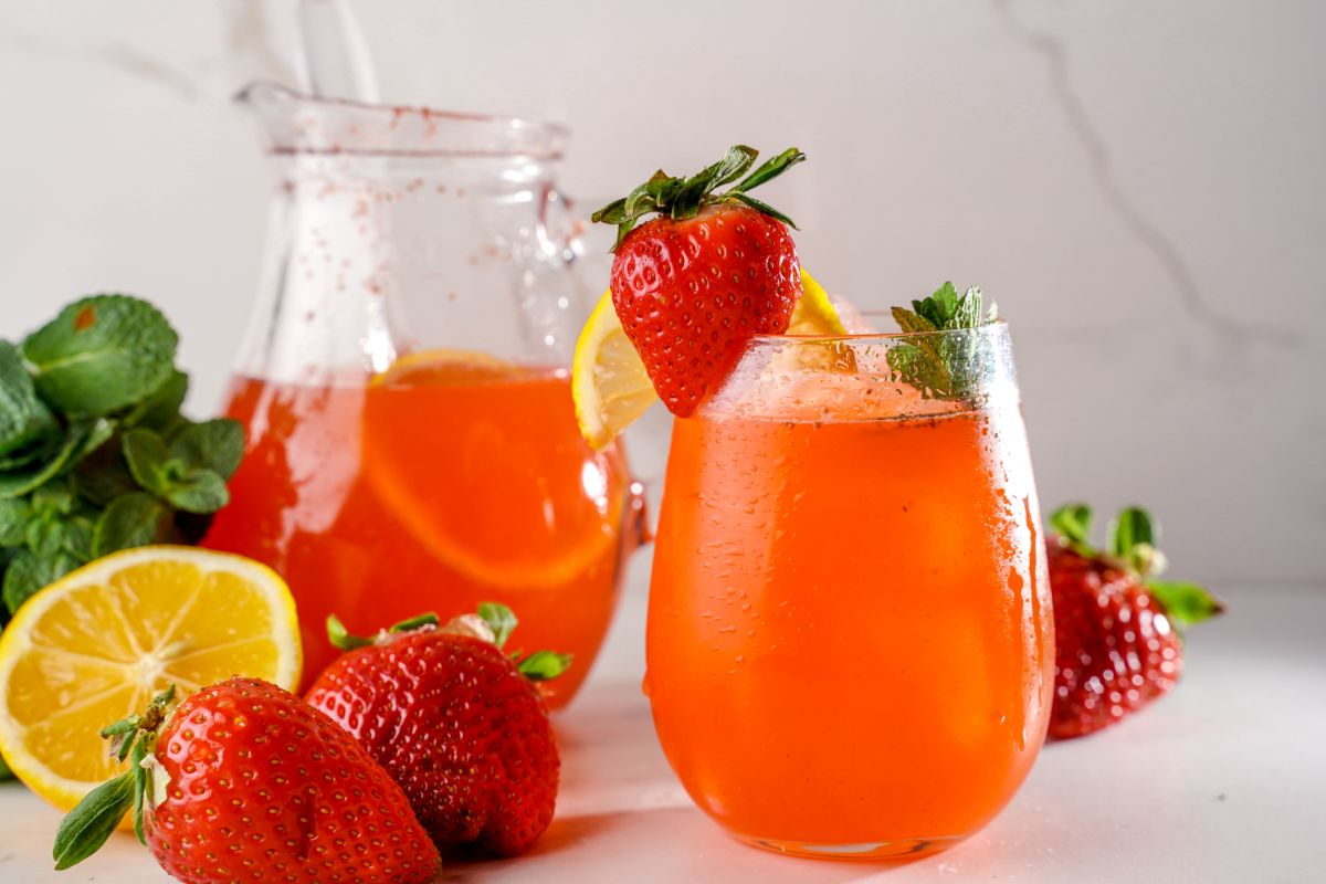 Strawberry lemonade in a glass pitcher and a glass cup with slices of lemon and strawberry.