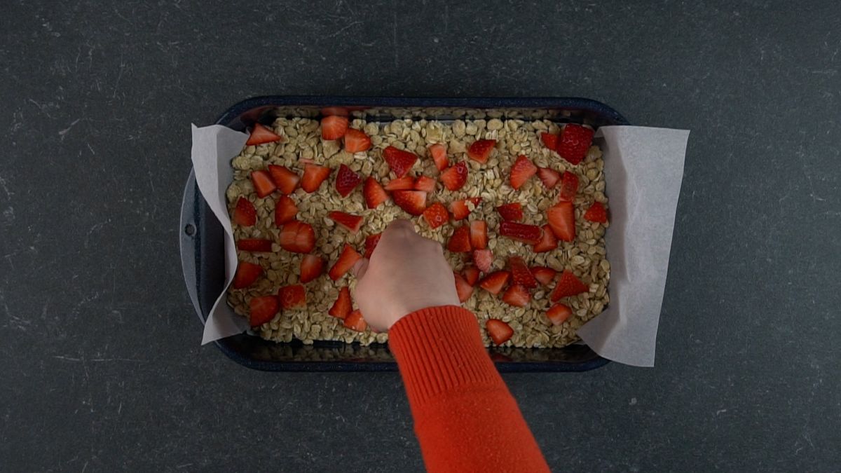 Hand putting sliced strawberries over mixture on a baking tray.