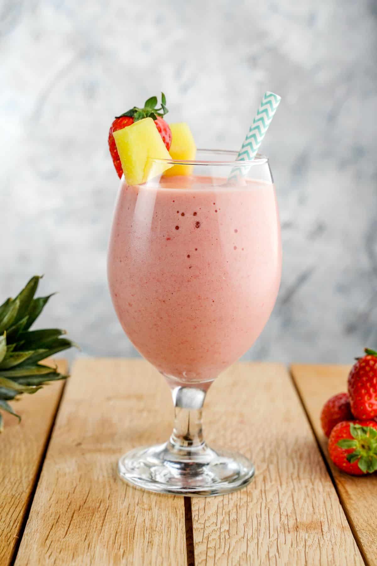 A close-up of a Strawberry Pineapple Smoothie in a tall glass with a straw nad piece of a strawberry and a pineapple.