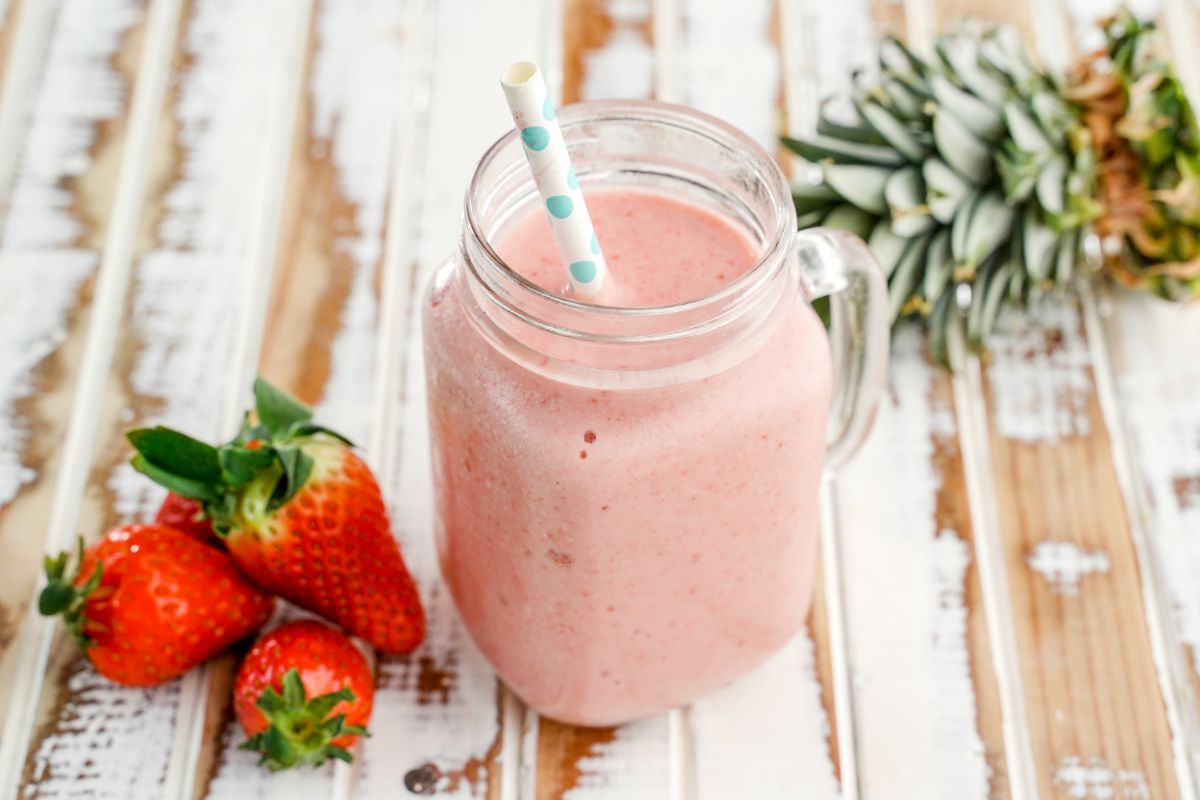 A glass jar of Strawberry Pineapple Smoothie with a straw on a wooden table with ripe strawberries and pineapple around.