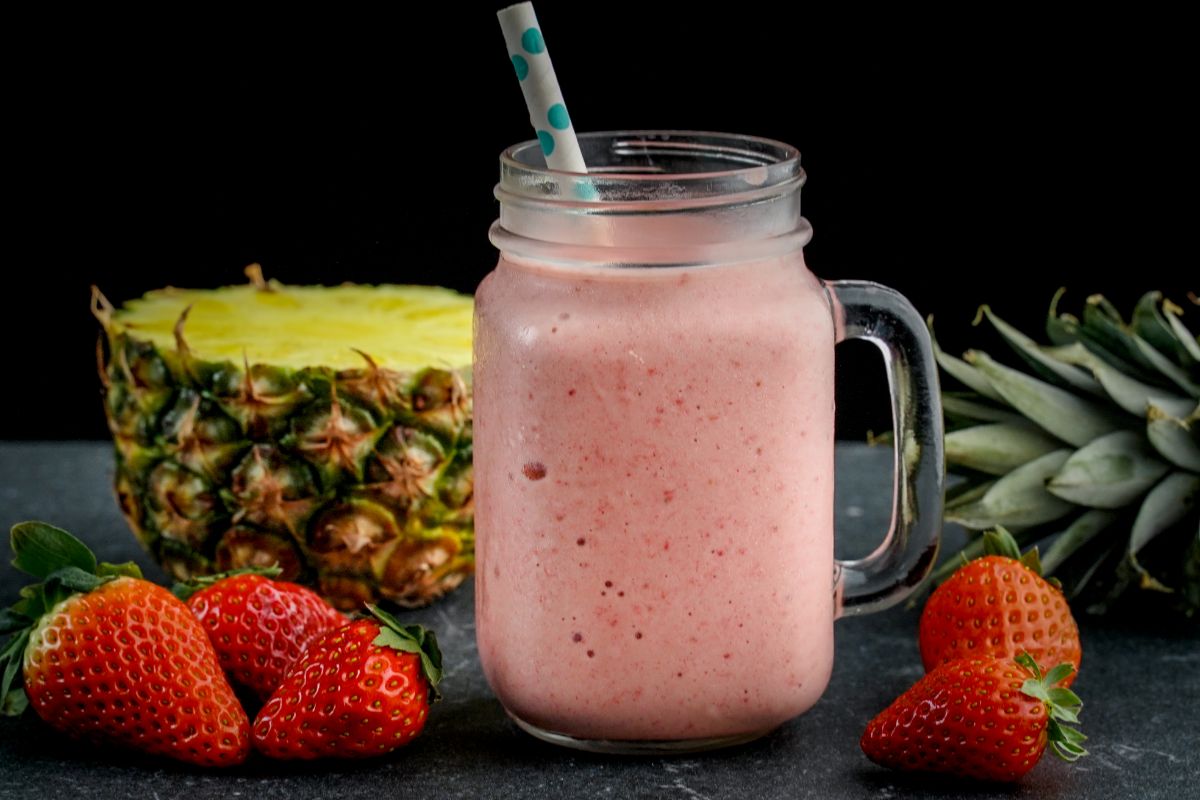 A glass jar of Strawberry Pineapple Smoothie with a straw on a table with ripe strawberries and pineapple around.