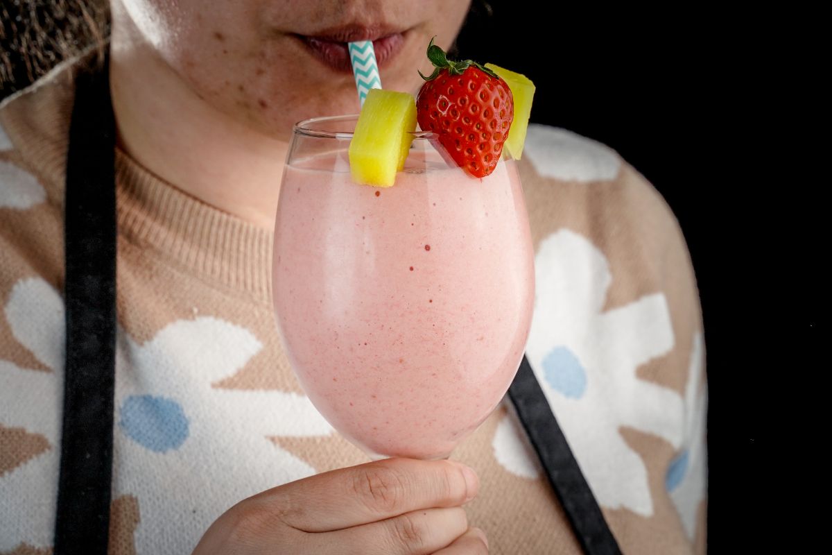 A woman sipping a Strawberry Pineapple Smoothie from a tall glass through a straw.
