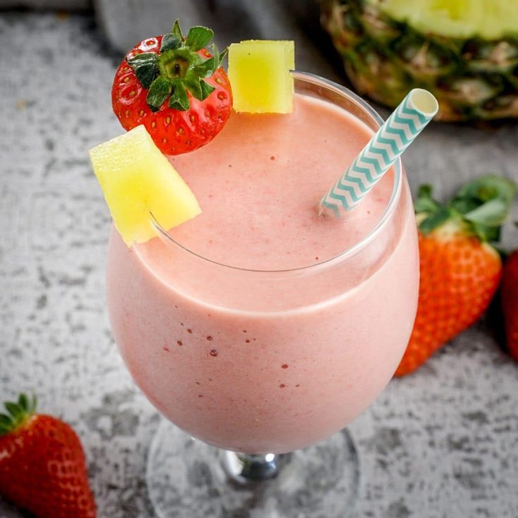 A close-up of a strawberry pineapple smoothie in a tall glass with a straw and slices of strawberry and pineapple.