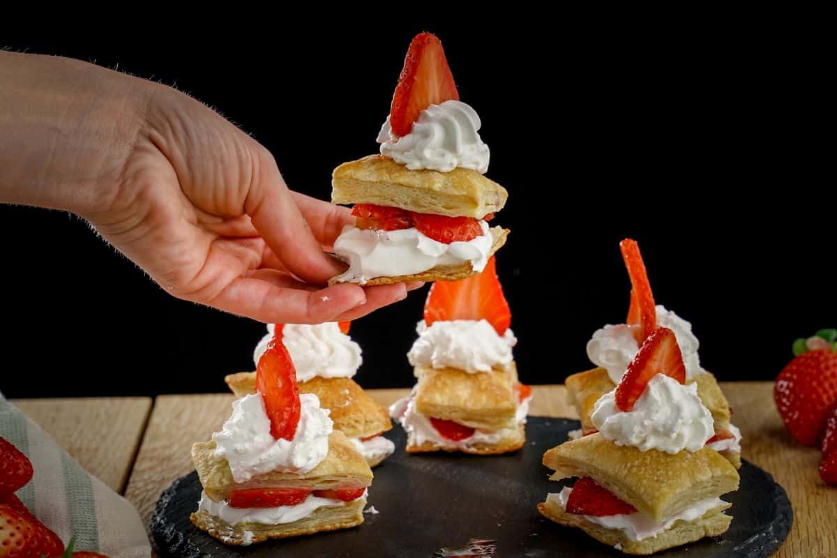 A hand holding a strawberry shortcake puff over a tray full of shortakce puffs.