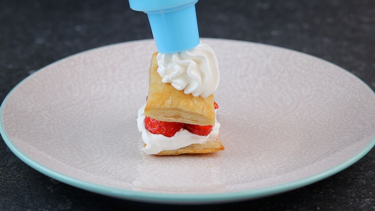 Putting a whipped cream on the top of a strawberry shortcake puff on a white plate.