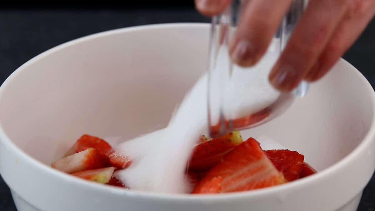 A close-up of pouring sugar in a white bowl with sliced strawberries.